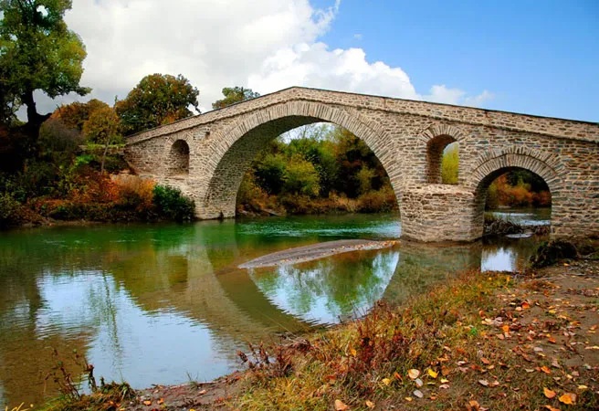 Το πέτρινο τοξωτό γεφύρι του Ζιάκα καθρεφτίζεται στα νερά του Βελονιά, ανάμεσα σε φθινοπωρινά δέντρα